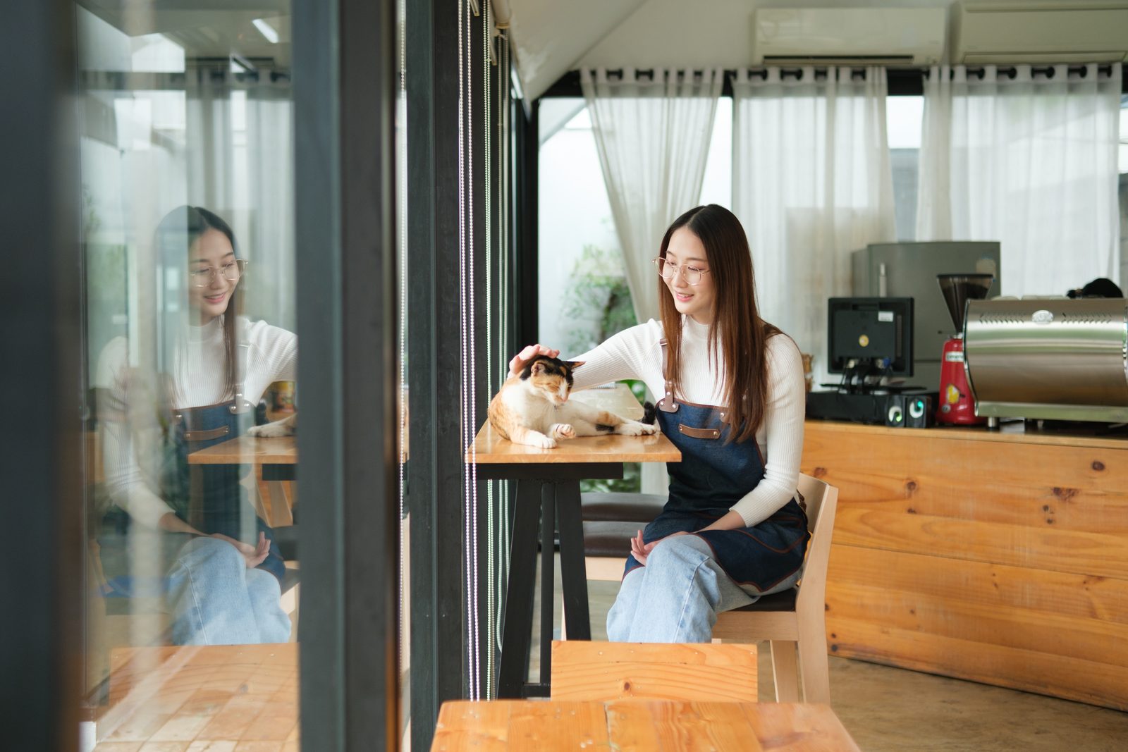 Woman in an apron sits at a wooden cafe table beside a calico cat, with an espresso machine and curtained windows behind