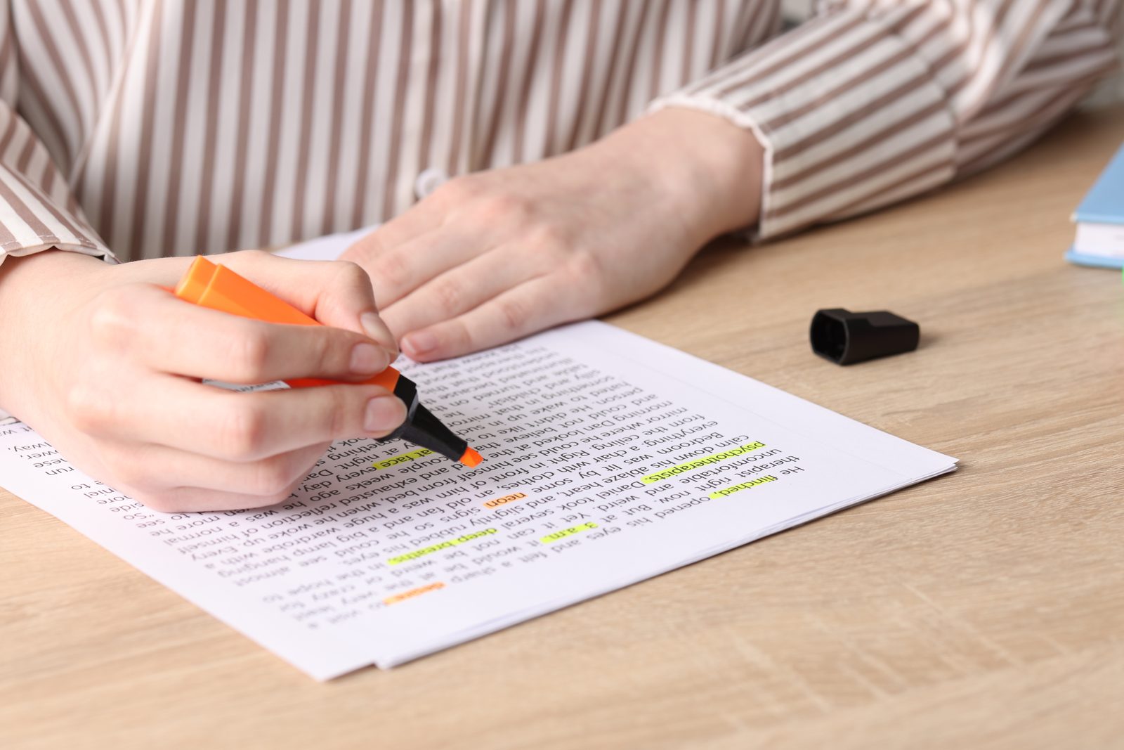 A hand holds an orange highlighter over a printed page of text on a wooden desk, with a few lines already marked.