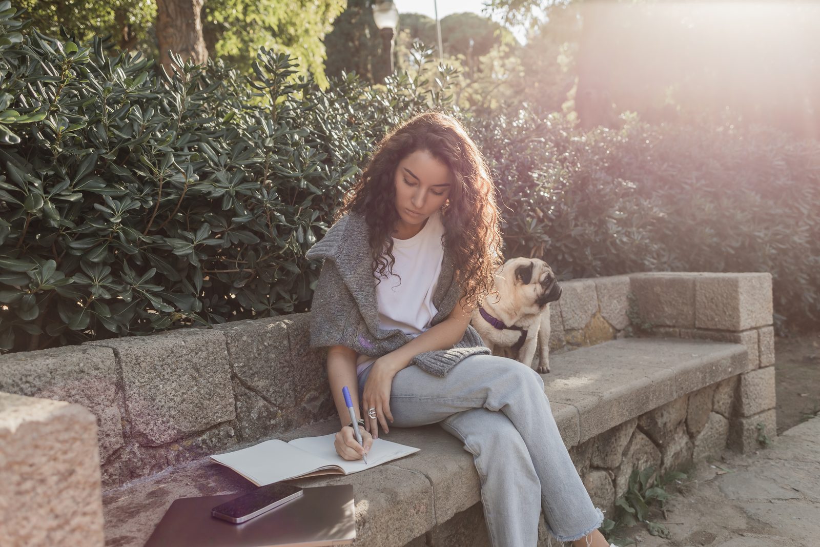 A curly haired woman writes in an open notebook on a stone garden bench in late sun while a pug sits on the bench beside her